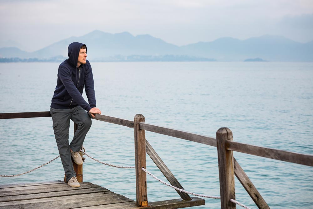 Man sitting on railing of wooden pier, sea and mountains view behind him