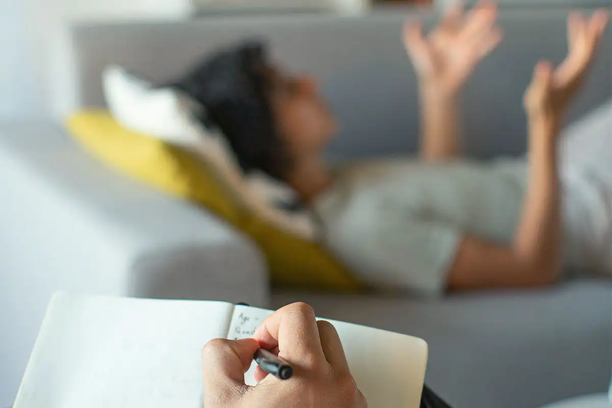 A woman engages in therapy, psychologist writing notes in a notebook while seated on a chair, during prescription drug addiction treatment